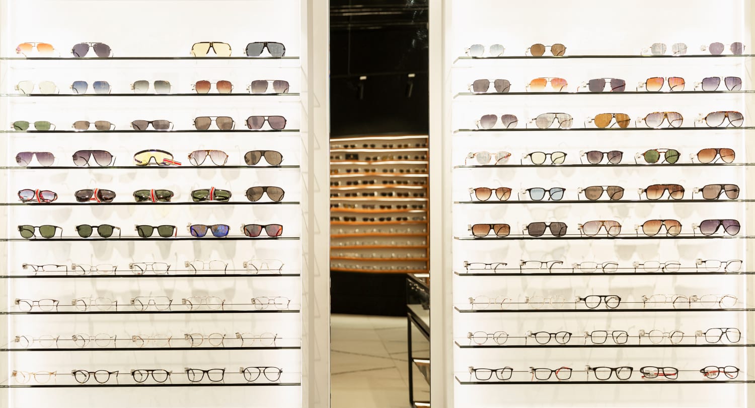 Eyewear store display with multiple rows of stylish eyeglasses and sunglasses neatly arranged on glass shelves, showcasing a wide product variety and inviting in-store atmosphere.